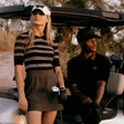 A woman and a man in golf attire pose in a golf cart outdoors. The woman wears a black and white striped polo shirt, a dark brown golf skirt, a white visor with a black logo, and a black glove while holding a club.