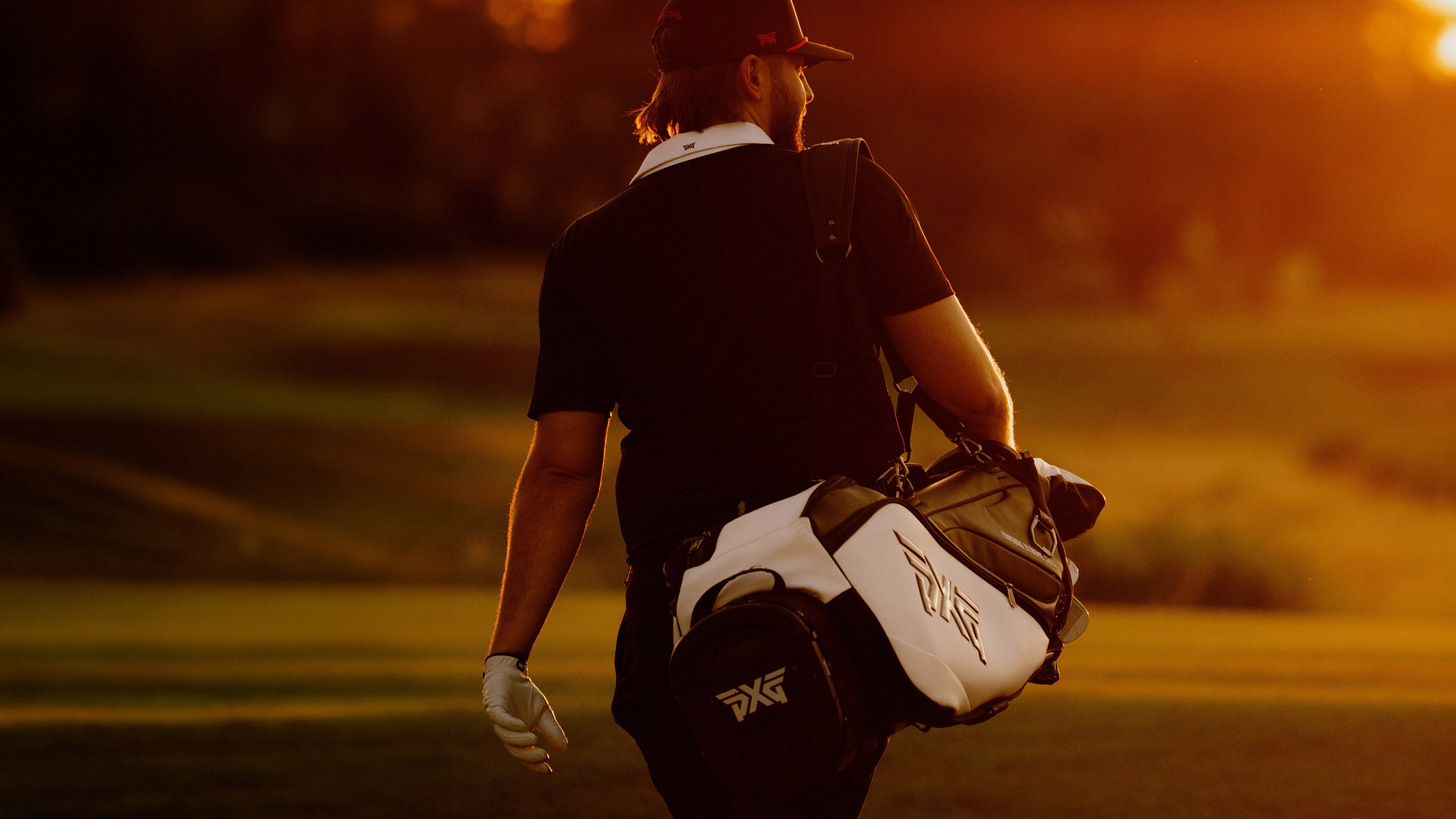 A golfer seen from behind on a golf course at sunset, carrying a white and black golf bag with a visible 
PXG logo.