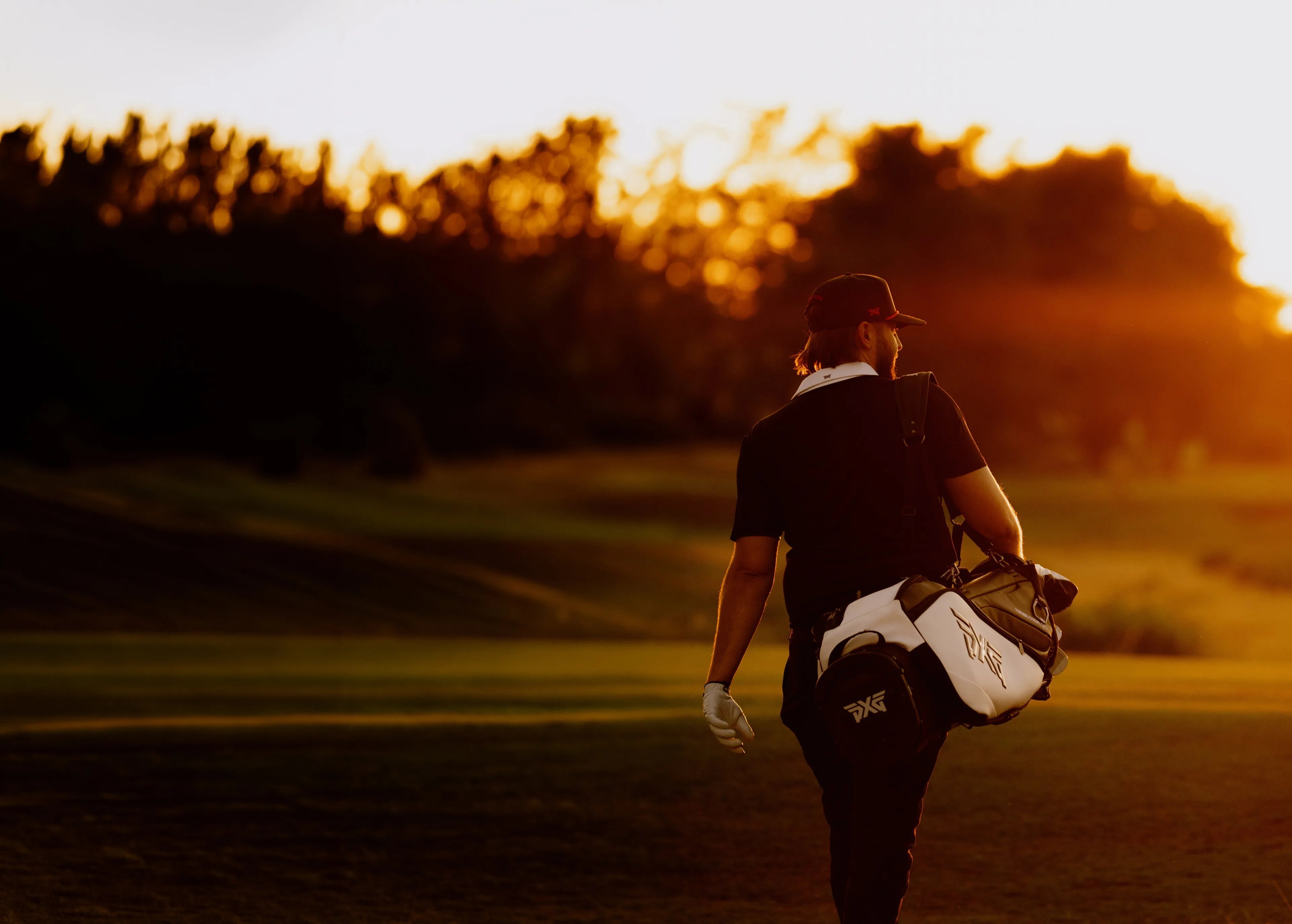 A golfer seen from behind walking on a golf course at sunset, carrying a white and black golf bag over his shoulder.